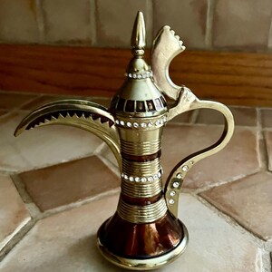 May include: A metallic gold-toned dallah coffee pot featuring a long curved spout, a pointed lid, and a handle. The body is decorated with horizontal bands of brown enamel and rows of small, clear, sparkling rhinestones along the neck and handle.