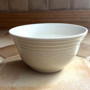 May include: A cream-colored ceramic bowl with a speckled finish. The bowl has a rounded shape with three horizontal, textured bands around the exterior. The interior is a bright white, contrasting with the exterior. This bowl is suitable for serving food.