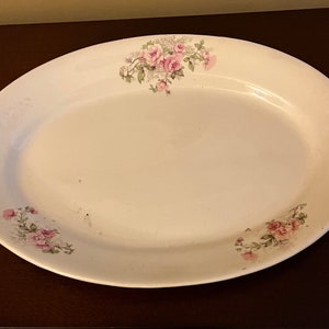 May include: Oval white serving platter with floral rose pattern in pink and green. The platter has a slightly raised edge and is suitable for serving food. The floral design is located on the edges of the platter.