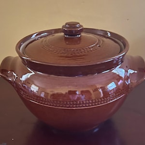 May include: A glossy brown ceramic pot with a matching lid and two side handles. The pot features a decorative band of small raised dots encircling the upper body, while the lid includes a central knob and a matching dotted pattern near the edge.