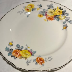 May include: A white ceramic plate with a gold rim and a floral design featuring yellow, orange, and pink roses. The roses are arranged in a circular pattern around the edge of the plate.