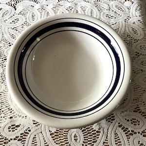 May include: A round white ceramic bowl featuring a wide dark blue band around the interior rim, accented by two thinner dark blue circular lines positioned just below the wider band, creating a simple and clean design for serving food.