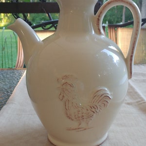 May include: A white ceramic pitcher with a rooster design. The pitcher has a brown handle and a spout.