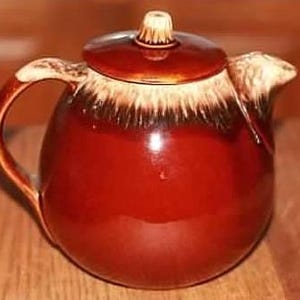 May include: A brown glazed ceramic teapot with a cream colored rim and lid. The teapot has a round body and a handle on the side.