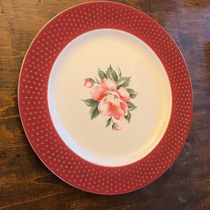 May include: A white ceramic dinner plate with a red and gold rim. The plate features a floral design with a pink flower and green leaves.