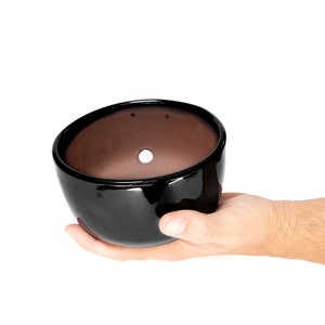 May include: A black ceramic pot with a drainage hole in the bottom. The pot is being held in a person's hand.