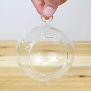May include: A clear glass sphere with a metal hanging loop. The sphere is empty and has a smooth, rounded surface.