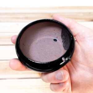 May include: A small, black ceramic bowl with a light purple interior. The bowl is slightly wider than it is tall and has a rounded shape.