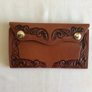 May include: A brown leather wallet with a decorative floral design. The wallet has a flap closure secured by two gold-colored snap buttons. The edges are stitched, and the overall design is embossed.
