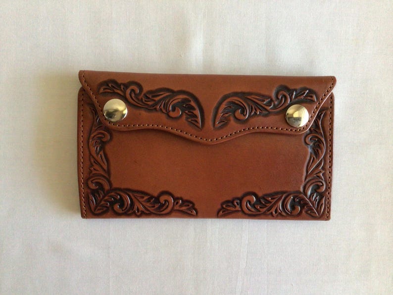 May include: A brown leather wallet with a decorative floral design. The wallet has a flap closure secured by two gold-colored snap buttons. The edges are stitched, and the overall design is embossed.
