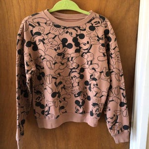 Op de afbeelding: Een sweatshirt met lange mouwen en een ronde hals in een stoffige roze kleur. Het sweatshirt is bedekt met een herhalend patroon van zwart-witte Mickey Mouse-stripfiguren. De mouwen en zoom hebben een geribbelde textuur.