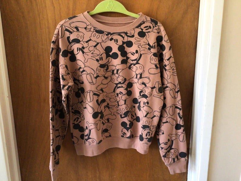Op de afbeelding: Een sweatshirt met lange mouwen en een ronde hals in een stoffige roze kleur. Het sweatshirt is bedekt met een herhalend patroon van zwart-witte Mickey Mouse-stripfiguren. De mouwen en zoom hebben een geribbelde textuur.