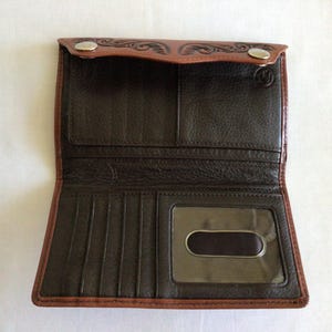 May include: Open brown leather wallet with a bi-fold design. The wallet features a dark brown interior with multiple card slots and a clear ID window. The exterior has a lighter brown trim and decorative embossed details.