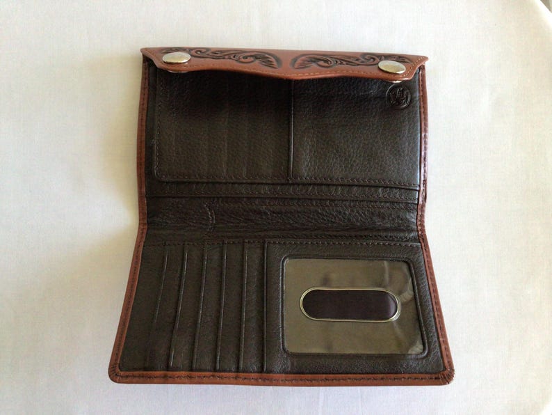 May include: Open brown leather wallet with a bi-fold design. The wallet features a dark brown interior with multiple card slots and a clear ID window. The exterior has a lighter brown trim and decorative embossed details.