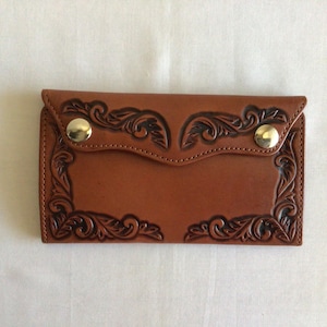 May include: A brown leather wallet with a decorative floral pattern embossed on the front. The wallet has a flap closure with two gold-colored snap closures. The edges are stitched, and the overall design is symmetrical.