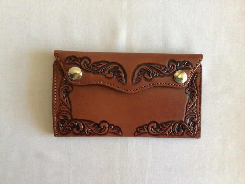 May include: A brown leather wallet with a decorative floral pattern embossed on the front. The wallet has a flap closure with two gold-colored snap closures. The edges are stitched, and the overall design is symmetrical.