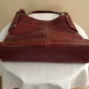 May include: A reddish-brown leather handbag with a crocodile-like texture. The bag has a shoulder strap and a rectangular base. The bag is sitting on a white surface.