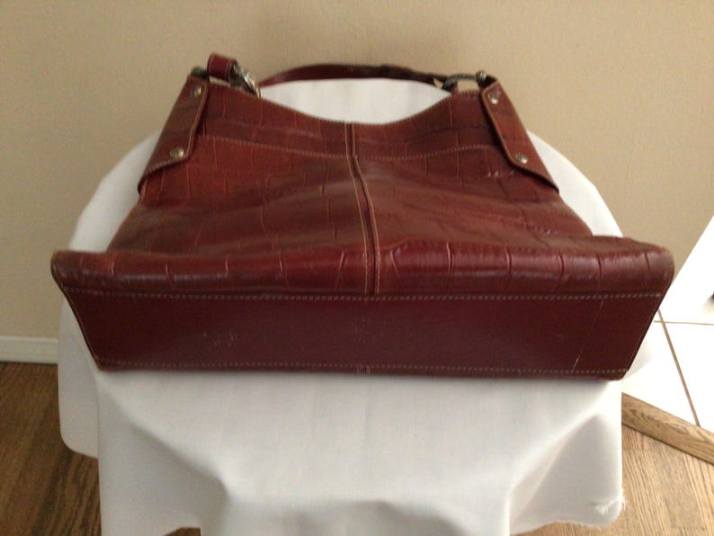 May include: A reddish-brown leather handbag with a crocodile-like texture. The bag has a shoulder strap and a rectangular base. The bag is sitting on a white surface.