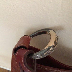May include: Close-up of a brown leather handbag with a silver-toned, oval-shaped handle. The handle has decorative scrollwork and shows signs of wear. The leather strap is stitched to the bag.