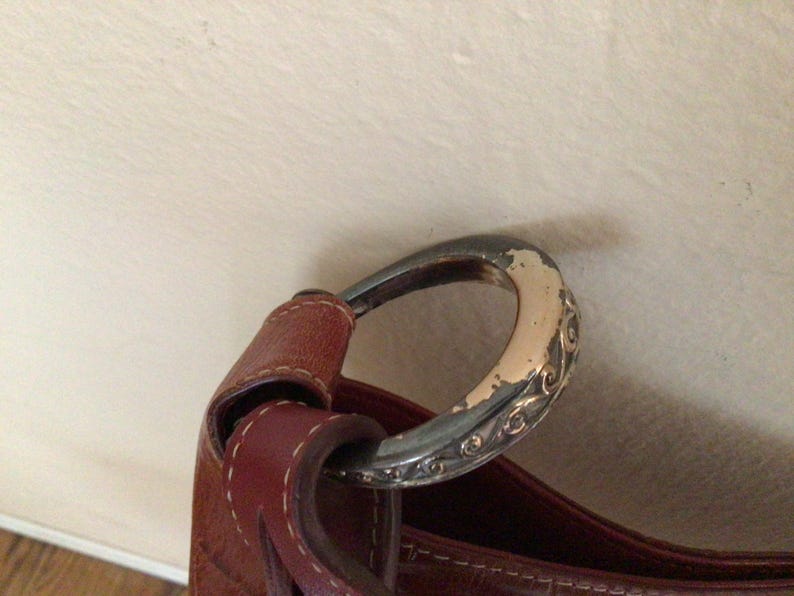 May include: Close-up of a brown leather handbag with a silver-toned, oval-shaped handle. The handle has decorative scrollwork and shows signs of wear. The leather strap is stitched to the bag.