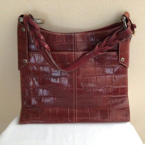 May include: A reddish-brown leather tote bag with a crocodile skin texture. The bag has a braided strap and a shoulder strap. Silver-toned hardware accents the bag's design. The bag is sitting on a white surface.