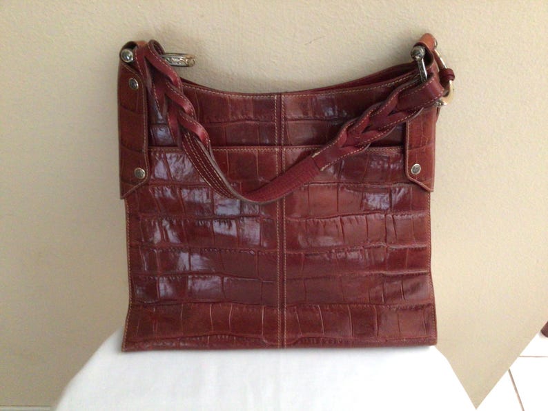 May include: A reddish-brown leather tote bag with a crocodile skin texture. The bag has a braided strap and a shoulder strap. Silver-toned hardware accents the bag's design. The bag is sitting on a white surface.
