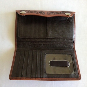 May include: Open brown leather wallet with a bi-fold design. The wallet features a dark brown interior with multiple card slots and a clear ID window. The exterior has a decorative embossed pattern and two snap closures.
