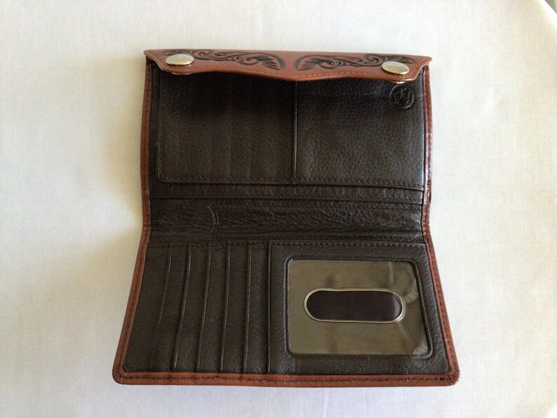 May include: Open brown leather wallet with a bi-fold design. The wallet features a dark brown interior with multiple card slots and a clear ID window. The exterior has a decorative embossed pattern and two snap closures.