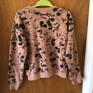 Op de afbeelding: Een lichtbruin sweatshirt met ronde hals en lange mouwen, bedekt met een herhalend patroon van zwarte Mickey Mouse-illustraties. Het sweatshirt hangt aan een groene hanger.