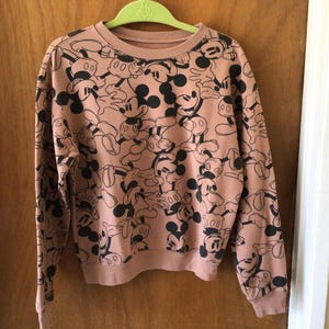Op de afbeelding: Een roze crewneck sweatshirt met lange mouwen, bedekt met een patroon van zwarte Mickey Mouse-illustraties. De manchetten en zoom zijn geribbeld. Het sweatshirt hangt aan een groene hanger.