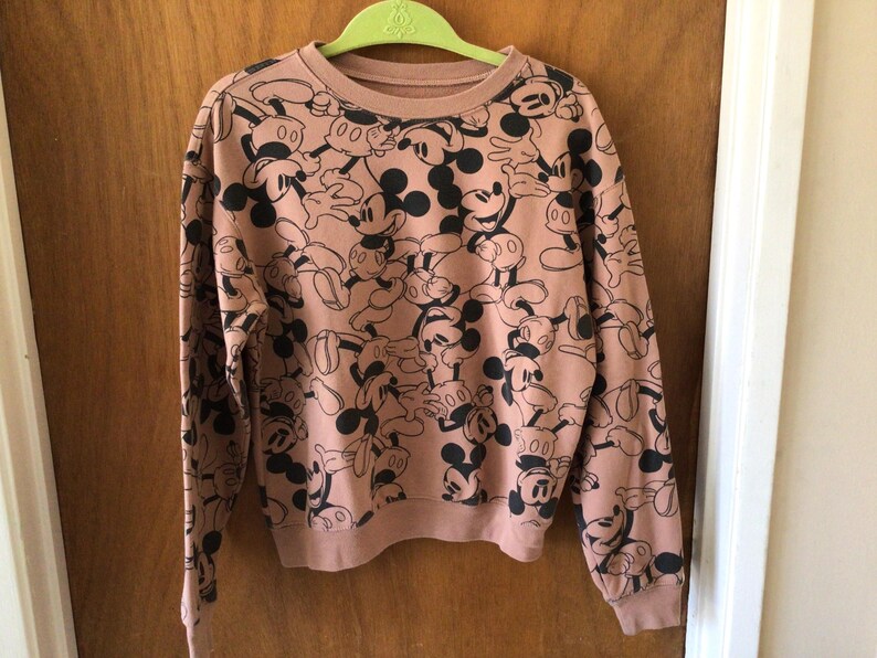 Op de afbeelding: Een roze crewneck sweatshirt met lange mouwen, bedekt met een patroon van zwarte Mickey Mouse-illustraties. De manchetten en zoom zijn geribbeld. Het sweatshirt hangt aan een groene hanger.