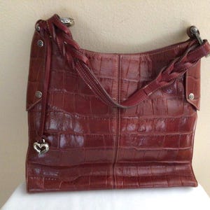 May include: A reddish-brown leather shoulder bag with a crocodile skin texture. The bag has a braided strap and a silver heart-shaped charm. The bag is sitting on a white surface.