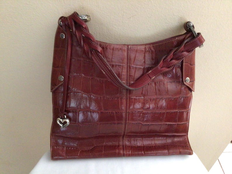 May include: A reddish-brown leather shoulder bag with a crocodile skin texture. The bag has a braided strap and a silver heart-shaped charm. The bag is sitting on a white surface.