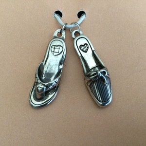 Pode incluir: Um par de pingentes de prata em forma de chinelo e sapato de salto alto. Ambos os pingentes t&ecirc;m um pequeno cora&ccedil;&atilde;o gravado.