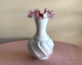 May include: A white ceramic vase with a ruffled, pink-trimmed rim. The vase has a bulbous base with a swirling design and a slender neck. The vase is sitting on a pink polka-dotted surface.