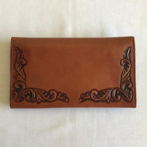 May include: A brown leather wallet with decorative corner accents. The wallet has a smooth surface with embossed floral designs in each corner. The leather is a warm, medium brown color.
