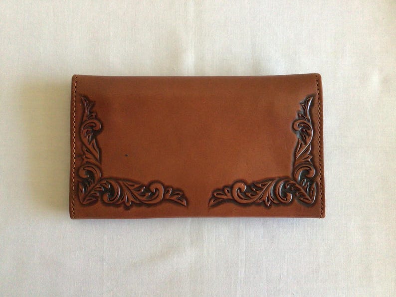 May include: A brown leather wallet with decorative corner accents. The wallet has a smooth surface with embossed floral designs in each corner. The leather is a warm, medium brown color.