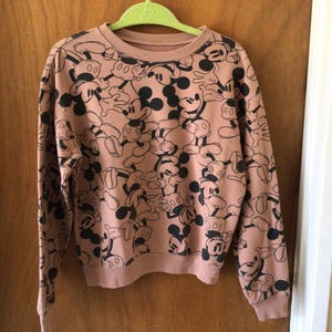 Op de afbeelding: Een sweatshirt met lange mouwen en een ronde hals in een stoffige roze kleur. Het sweatshirt is bedekt met een herhalend patroon van zwart-witte Mickey Mouse-stripfiguren. De manchetten en zoom zijn geribbeld.