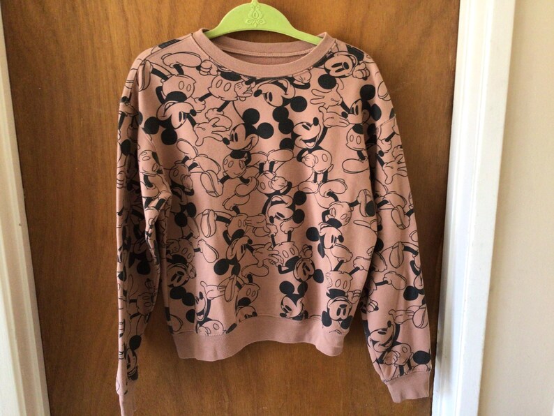 Op de afbeelding: Een sweatshirt met lange mouwen en een ronde hals in een stoffige roze kleur. Het sweatshirt is bedekt met een herhalend patroon van zwart-witte Mickey Mouse-stripfiguren. De manchetten en zoom zijn geribbeld.