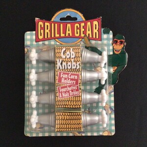 May include: A package of "Grilla Gear" cob knobs, fun corn holders. The package features six silver corn holders with white handles. The packaging has a green and white checkered pattern and a cartoon monkey wearing sunglasses.