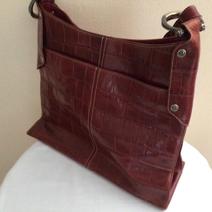 May include: A reddish-brown shoulder bag with a crocodile-like texture. The bag has a rectangular shape with a front pocket and a metal ring handle. The bag is sitting on a white surface.