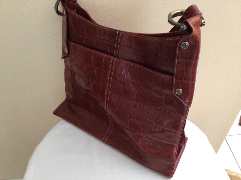 May include: A reddish-brown shoulder bag with a crocodile-like texture. The bag has a rectangular shape with a front pocket and a metal ring handle. The bag is sitting on a white surface.