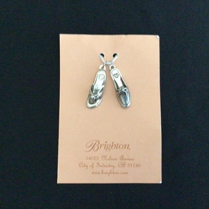 Pode incluir: Dois pingentes de prata em forma de sand&aacute;lias, um com um design simples e o outro com um design mais detalhado. Os pingentes est&atilde;o em um cart&atilde;o branco com o texto "Brighton 14022 Nelson Avenue City of Industry, CA 91746 www.brighton.com"