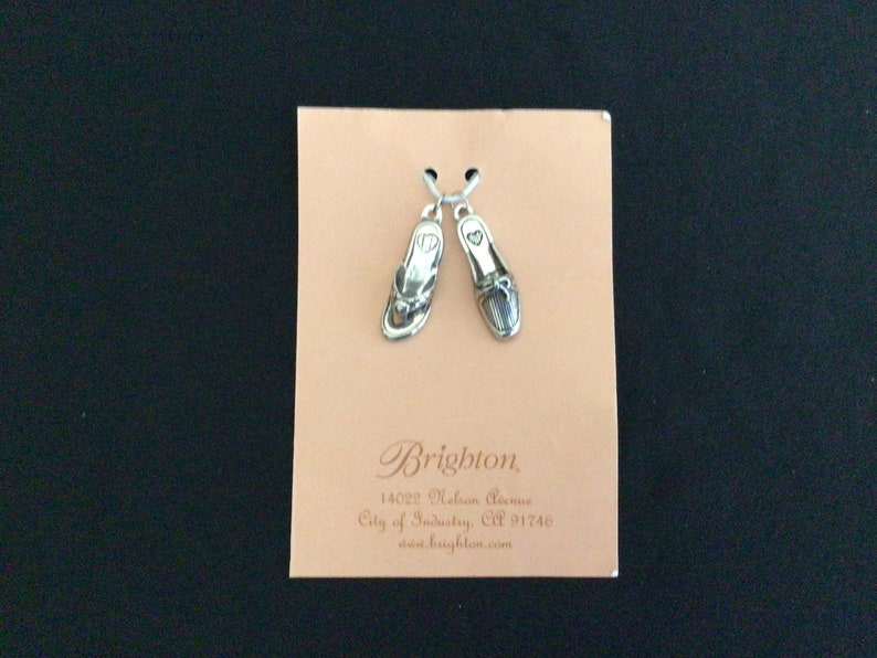 Pode incluir: Dois pingentes de prata em forma de sand&aacute;lias, um com um design simples e o outro com um design mais detalhado. Os pingentes est&atilde;o em um cart&atilde;o branco com o texto "Brighton 14022 Nelson Avenue City of Industry, CA 91746 www.brighton.com"