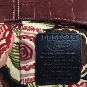May include: Close-up of a handbag interior with a floral pattern in shades of red, pink, green, and cream. The top edge is a dark brown, textured leather. A black leather tag displays the Brighton logo and text.