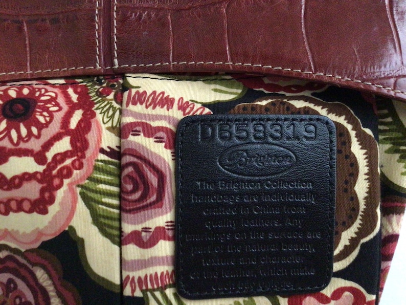 May include: Close-up of a handbag interior with a floral pattern in shades of red, pink, green, and cream. The top edge is a dark brown, textured leather. A black leather tag displays the Brighton logo and text.