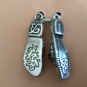 Pode incluir: Dois pingentes de prata em forma de sapato com desenhos intrincados. Os pingentes s&atilde;o conectados por um anel de prata.
