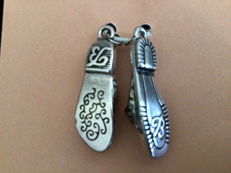 Pode incluir: Dois pingentes de prata em forma de sapato com desenhos intrincados. Os pingentes s&atilde;o conectados por um anel de prata.