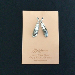 Pode incluir: Um par de pulseiras de prata com um amuleto de chinelo e um amuleto de mocassim. Os amuletos est&atilde;o em um cart&atilde;o com o texto "Brighton, 14022 Nelson Avenue, City of Industry, CA 91746, www.brighton.com".