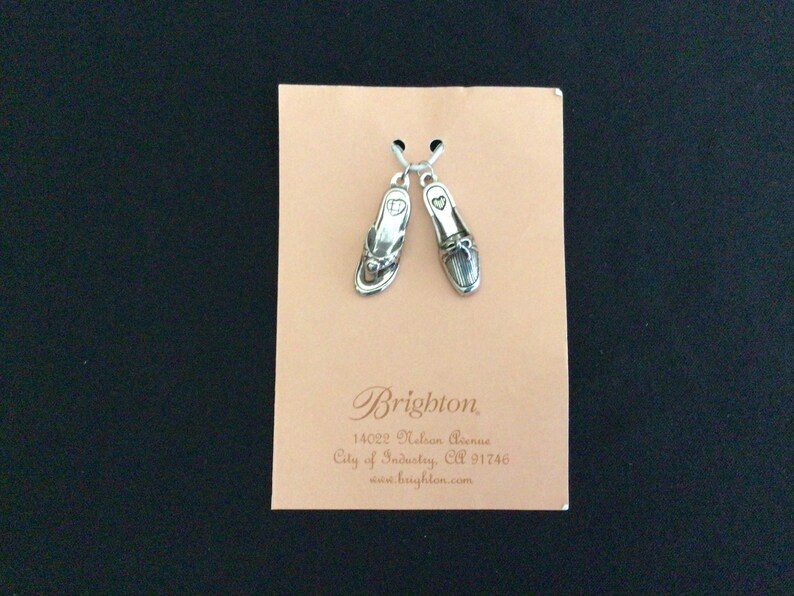 Pode incluir: Um par de pulseiras de prata com um amuleto de chinelo e um amuleto de mocassim. Os amuletos est&atilde;o em um cart&atilde;o com o texto "Brighton, 14022 Nelson Avenue, City of Industry, CA 91746, www.brighton.com".